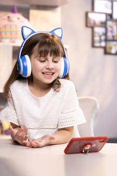 Happy cute small caucasian kid girl vlogger waving hand talking to camera at home recording vlog for social media blog, video conference calling virtual friend having online meeting sitting in chair.