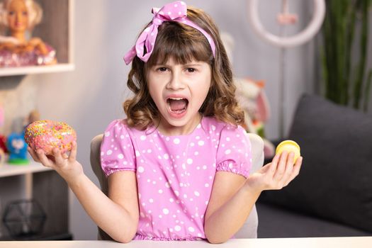 Cute little girl in pink dress holding donut and macaron in hands. Cute adolescent school girl plays with sweet donut macaroon doing happy fun face expressions on background. Funny concept with sweets