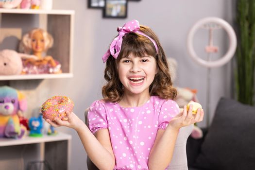Portrait of child girl choosing between two donuts in home room. Cheerful school girl playing with cakes indoors. Funny teenager girl having fun with colorful donuts at modern home.