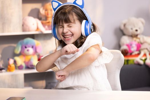 Happy cute small caucasian kid girl in headphones dancing and singing having fun in apartment. People, lifestyle and joy concept.