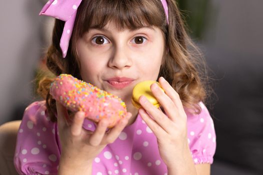 Adolescent school girl plays with sweet donuts doing happy fun face expressions on background. Cute little girl in pink dress holding macaron and donut in hands by the face. Funny concept with sweets.