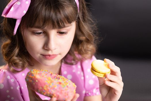 Close up of attractive school girl on home background, licks her lips, sniffing donut and yellow macaron, closes her eyes with pleasure. Dessert person, sweet tooth, gourmet. Feeling good, delicious.