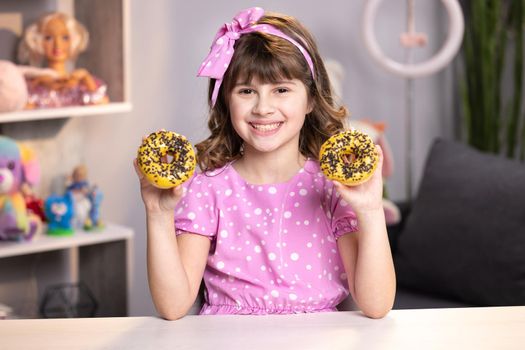 Portrait of sweet girl choosing between two donuts in home room. Cheerful school girl playing with cakes indoors. Funny teenager girl having fun with colorful donuts at modern home.