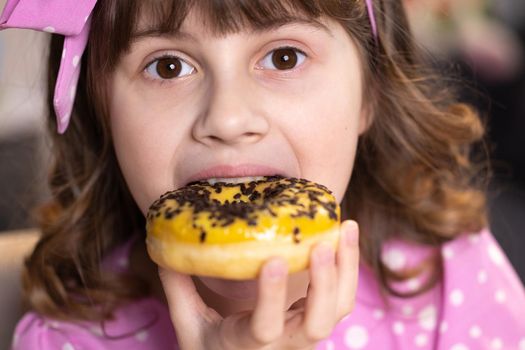 Close up school girl eating donut while sitting at table at home. Portrait of sweet girl choosing donut indoors. Funny teenager girl having fun with yellow donuts at modern home.