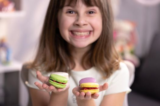 Smiling cute school kid girl holds four tasty cookies in her hands and look at camera. Traditional French multicolored macaroon. Food concept.