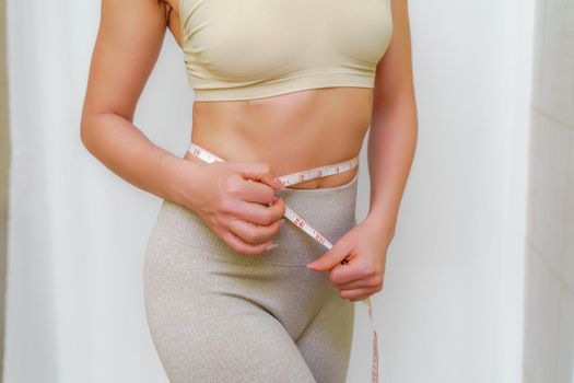 Cropped view of slim woman measuring waist with tape measure at home, close up. An unrecognizable European woman checks the result of a weight loss diet or liposuction indoors. Healthy lifestyle