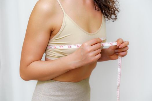 Cropped view of slim woman measuring breasts with tape measure at home, close up. Unrecognizable European woman checks the result of a weight loss diet or liposuction indoors. Healthy lifestyle..