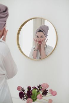 young woman with a towel on head looking in the mirror and apply cream on face skin.
