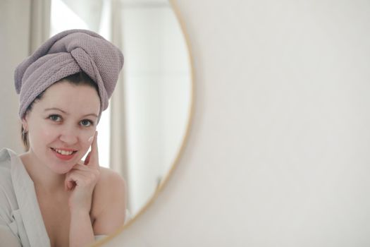 young woman with a towel on head looking in the mirror and apply cream on face skin.