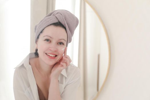 young woman with a towel on head looking in the mirror and apply cream on face skin.