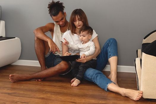 A happy, modern, multiracial family with a little boy in casual clothes sits against a gray wall in a room, looks into a digital tablet. copy space