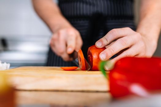 detail of the hands of a young man, dressed in a black polo shirt and a dark striped apron, cutting vegetables with a knife on a wooden board in a professional kitchen, on a table full of food. Cold and clean environment, background of professional aluminium tables and cooking fires of a restaurant.