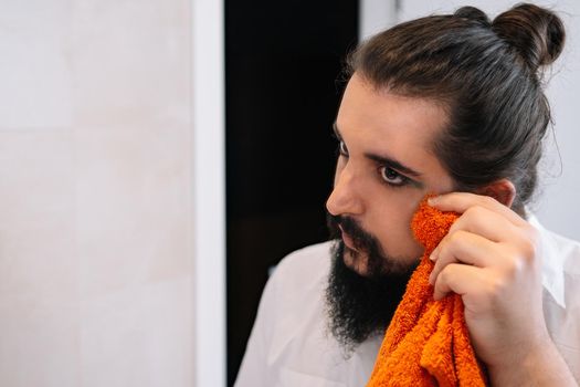 Smiling young woman touching up her eye make-up with a towel. Daily cosmetic routine of non-binary gender. Young woman with beard and long hair tied back applying make-up in the mirror. Image reflected in a mirror. Smiling young woman applying make-up in her bathroom. Trans with beautiful eyelashes.