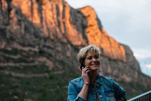 Portrait of an adult blonde short haired modern woman, smiling and happy, dressed in a casual way, with blue denim jacket and long coloured skirt, talking on mobile phone with her family, telling how she enjoys her holidays. Warm and calm atmosphere with sunset light, background of mountains and forests.