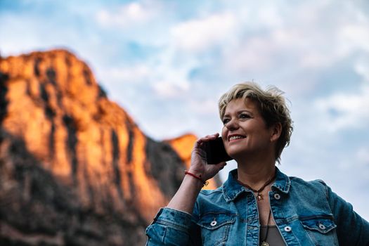 Portrait of an adult blonde short haired modern woman, smiling and happy, dressed in a casual way, with blue denim jacket and long coloured skirt, talking on mobile phone with her family, telling how she enjoys her holidays. Warm and calm atmosphere with sunset light, background of mountains and forests, blue sky with big clouds.