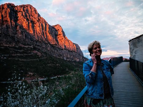 Portrait of an adult blonde short haired modern woman, smiling and happy, casually dressed, wearing blue denim jacket, long coloured skirt, on holiday walking and talking on the phone with her family, tells how she enjoys her getaway. Warm and calm atmosphere with sunset light, background of mountains and forests, blue sky with big clouds.