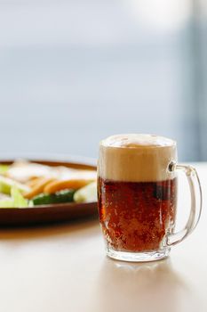 Beer cup standing near appetizers plate. Dark beer with thick high foam. Standing on smooth wooden surface of restaurant's or pub table. Looking mouthwatering. Cold and fresh. For friends company.