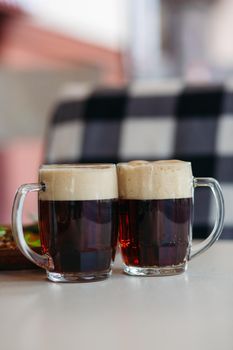 Two big glass cups full of foamy cold beer standing on pub's table in front of black and white checkered spread. Dark, fresh bear. Best drink for summer heat. Good for friends company.