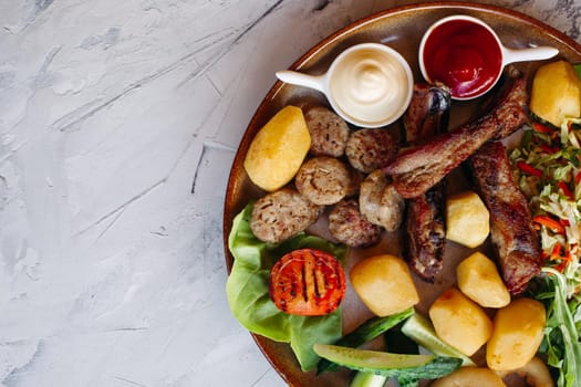 Top photo of clay plate full of tasty appetizers for beer. vegetable salad, boiled potatoes, grilled chicken legs, roasted goldy onion and sausages, cucmbers, tomatoes and sauces - ketchup and mayo.