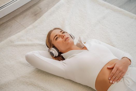 Top view portrait of relaxed woman listening to music with headphones lying on carpet at home. She is dressed in a White tracksuit