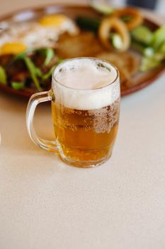Vertical shot of foamy cold beer cup standing near appetizers plate. Light fresh beer with thick high foam. Standing on smooth wooden surface of restaurant's or pub table. Looking mouthwatering.