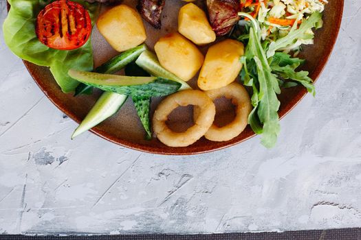 Top view of clay plate full of appetizers for beer including vegetable salad, boiled potatoes, grilled chicken legs, roasted goldy onion and sausages, cucmbers, tomatoes and sauces - ketchup and mayo.