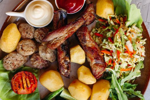 Horizontal photo of tasty food laying on clay plate. Delicious set of vegetable salad, boiled potatoes, grilled chicken legs, roasted onion, sausages, cucmbers, tomatoes and sauces - ketchup and mayo.