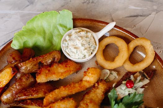 Delicious appetizers for beer and light alcohol drinks laying on clay plate decorated with salad leaves and cheese: crispy chicken nuggets, canapes with herring and cherry tomatoes, garlic sauce.