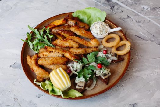 Clay plate full of appetizers including goldy chicken nuggets with chrispy crust, delicious canapes with herring and cherry tomatoes, served with garlic sauce, decorated with salad leaves and cheese.