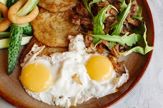 Delicious potato puncakes lying on brown clay plate with fried eggs. Fresh green cucumbers, fried golden onion and crusty meat, served with salad leaves. Tasty mouthwatering morning's meal.
