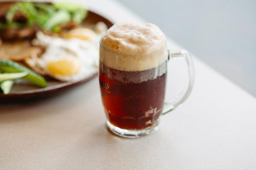 Beer cup standing near appetizers plate. Dark beer with thick high foam. Standing on smooth wooden surface of restaurant's or pub table. Looking mouthwatering. Cold and fresh. For friends company.