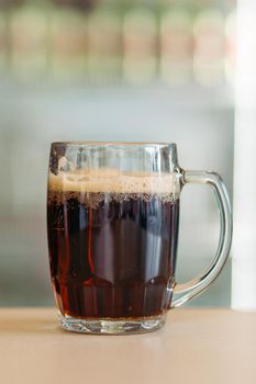 Photo of glass beer cup standing on smooth wooden surface. Cold dark summer drink for day heat. Fresh with thick high foam. Light blurred background. Concept of light alcohol drinks shooting.