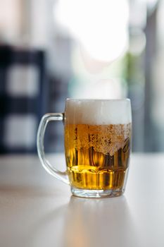Horizontal photo of glass cup full of light fresh beer standing on smooth wooden surface. Cold summer drink for day heat. Fresh with thick high foam. Blurred background. Concept of drinks shooting.