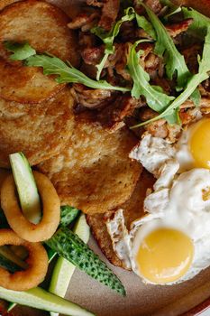 Delicious potato puncakes lying on brown clay plate with fried eggs. Fresh green cucumbers, fried golden onion and crusty meat, served with salad leaves. Tasty mouthwatering morning's meal.