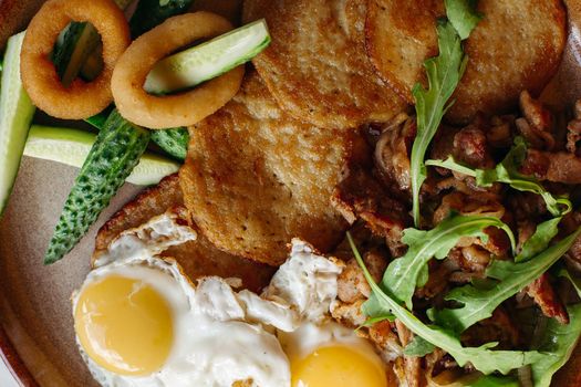 Delicious potato puncakes lying on brown clay plate with fried eggs. Fresh green cucumbers, fried golden onion and crusty meat, served with salad leaves. Tasty mouthwatering morning's meal.