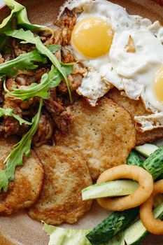 Delicious potato puncakes lying on brown clay plate with fried eggs. Fresh green cucumbers, fried golden onion and crusty meat, served with salad leaves. Tasty mouthwatering morning's meal.