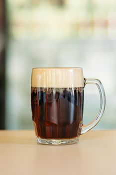 Photo of glass beer cup standing on smooth wooden surface. Cold dark summer drink for day heat. Fresh with thick high foam. Light blurred background. Concept of light alcohol drinks shooting.