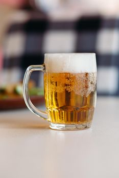 Horizontal photo of glass cup full of light fresh beer standing on smooth wooden surface. Cold summer drink for day heat. Fresh with thick high foam. Blurred background. Concept of drinks shooting.