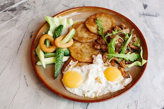 Delicious potato puncakes served with fried eggs laying on glancy clay plate including fresh green cucumbers, fried golden onion and crusty meat, served with salad leaves. Looking tasty. White table.