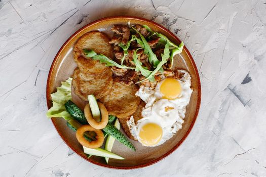 Delicious potato puncakes served with fried eggs laying on glancy clay plate including fresh green cucumbers, fried golden onion and crusty meat, served with salad leaves. Looking tasty. White table.