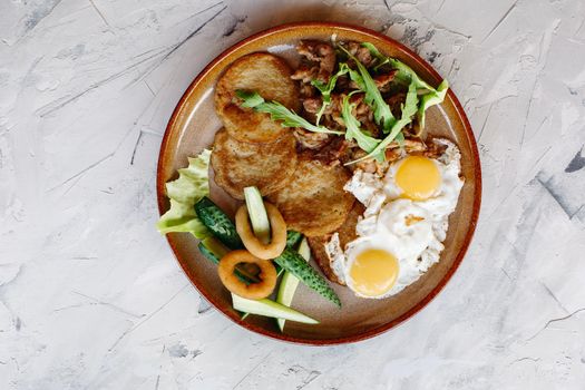 Delicious potato puncakes served with fried eggs laying on glancy clay plate including fresh green cucumbers, fried golden onion and crusty meat, served with salad leaves. Looking tasty. White table.