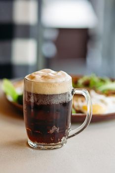 Beer cup standing near appetizers plate. Dark beer with thick high foam. Standing on smooth wooden surface of restaurant's or pub table. Looking mouthwatering. Cold and fresh. For friends company.