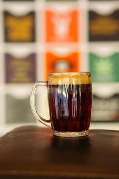 Photo of glass beer cup standing on smooth wooden surface. Cold dark summer drink for day heat. Fresh with thick high foam. Light blurred background. Concept of light alcohol drinks shooting.