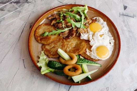 Delicious potato puncakes served with fried eggs laying on glancy clay plate including fresh green cucumbers, fried golden onion and crusty meat, served with salad leaves. Looking tasty. White table.