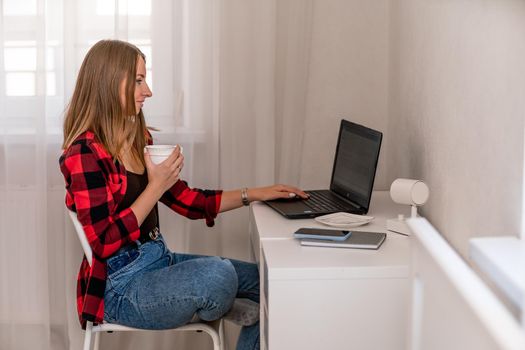 European professional woman sitting with laptop at home office desk, positive woman studying while working on PC. She is wearing a red plaid shirt and jeans