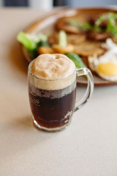 Beer cup standing near appetizers plate. Dark beer with thick high foam. Standing on smooth wooden surface of restaurant's or pub table. Looking mouthwatering. Cold and fresh. For friends company.