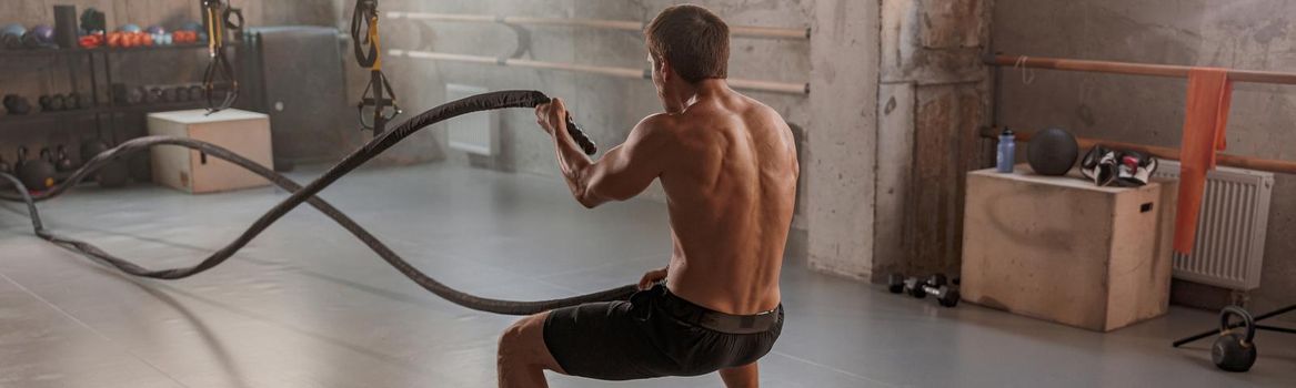 Back view of strong shirtless muscular man with battle rope doing exercise in functional training fitness gym