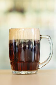 Photo of glass beer cup standing on smooth wooden surface. Cold dark summer drink for day heat. Fresh with thick high foam. Light blurred background. Concept of light alcohol drinks shooting.