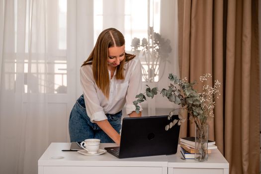 European professional woman is sitting with a laptop at a table in a home office, a positive woman is studying while working on a PC. She is wearing a beige jacket and jeans and is on the phone