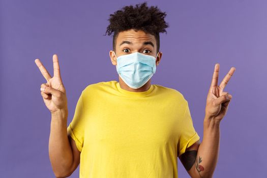 Medicine, covid19, coronavirus and people concept. Upbeat, hispanic man with afro haircut, wear medical face mask and show peace signs, staying positive, talking video-call while social distancing.